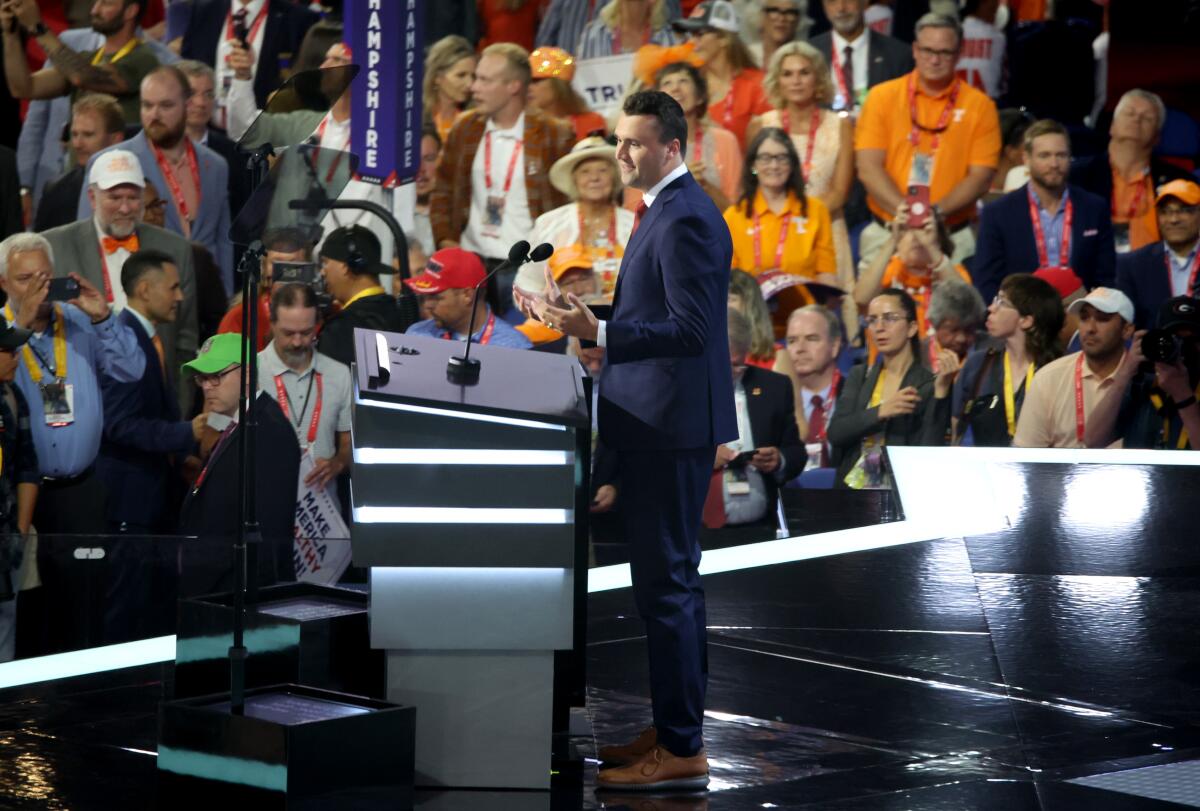 A man in a suit stands at a lectern on a stage in front of several people in an audience.