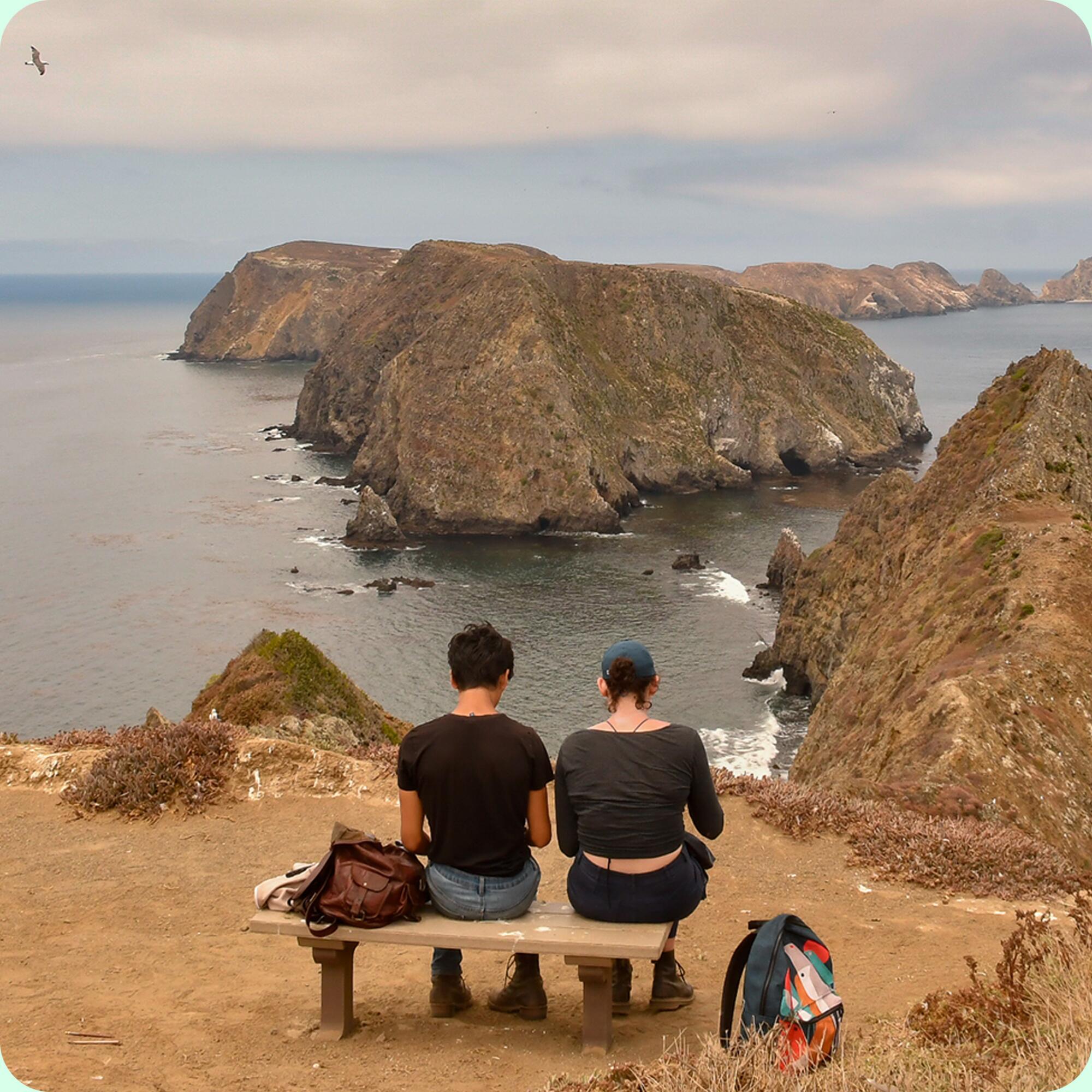 Two people sit on a bench, backs to the camera, looking toward rock formations in the ocean