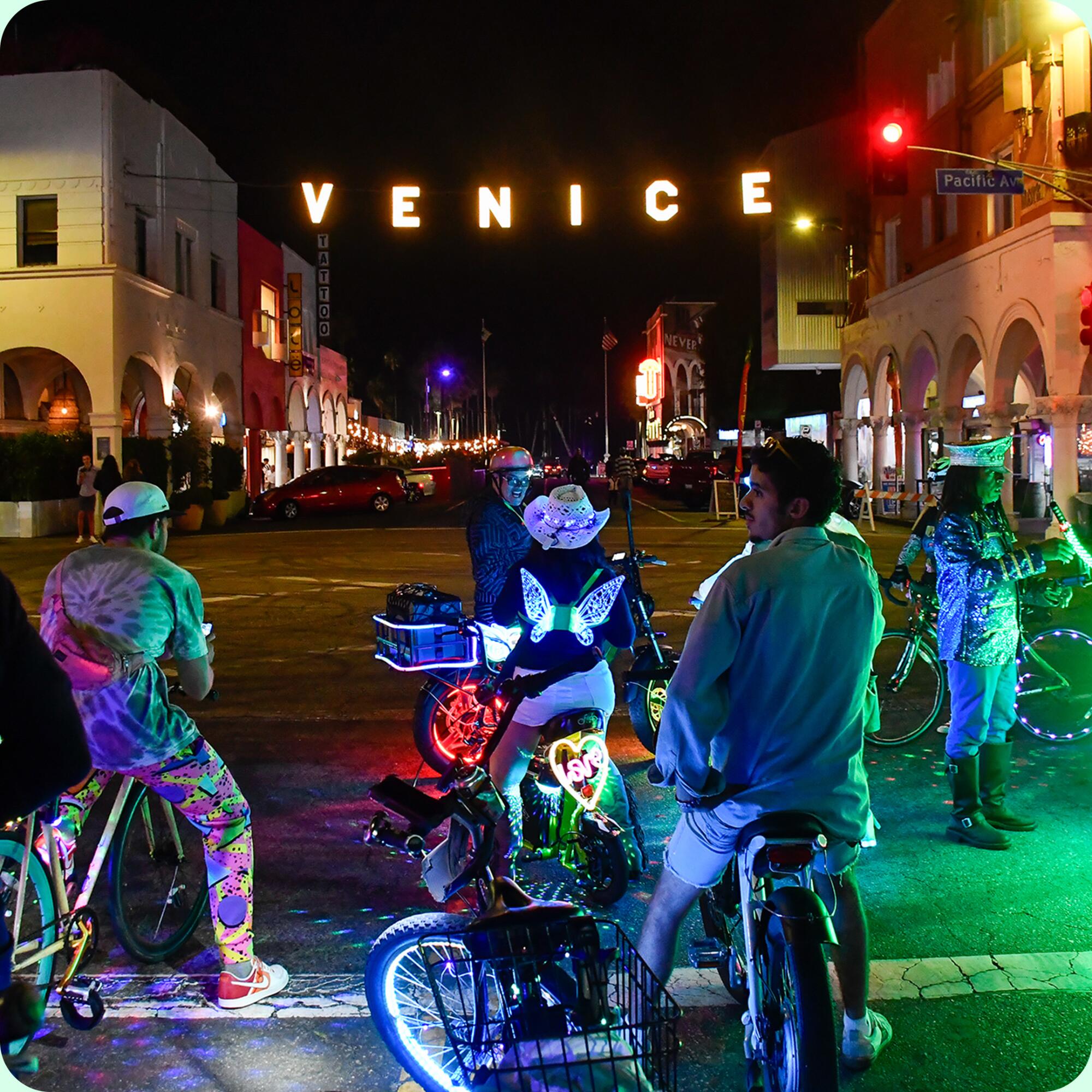 Bike riders at night stand looking toward the illuminated Venice sign that stretches across a city street. 