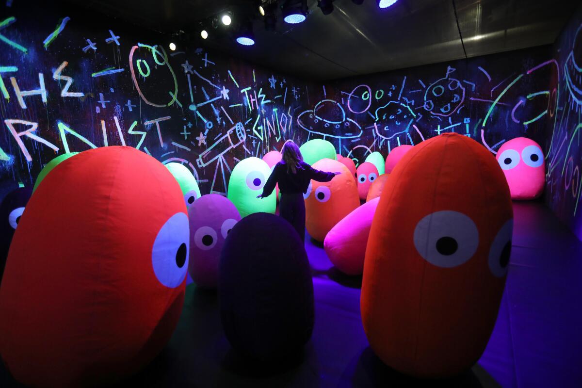 A person stands among colorful blobs with one or two eyes, in a room with child-like drawings on the walls