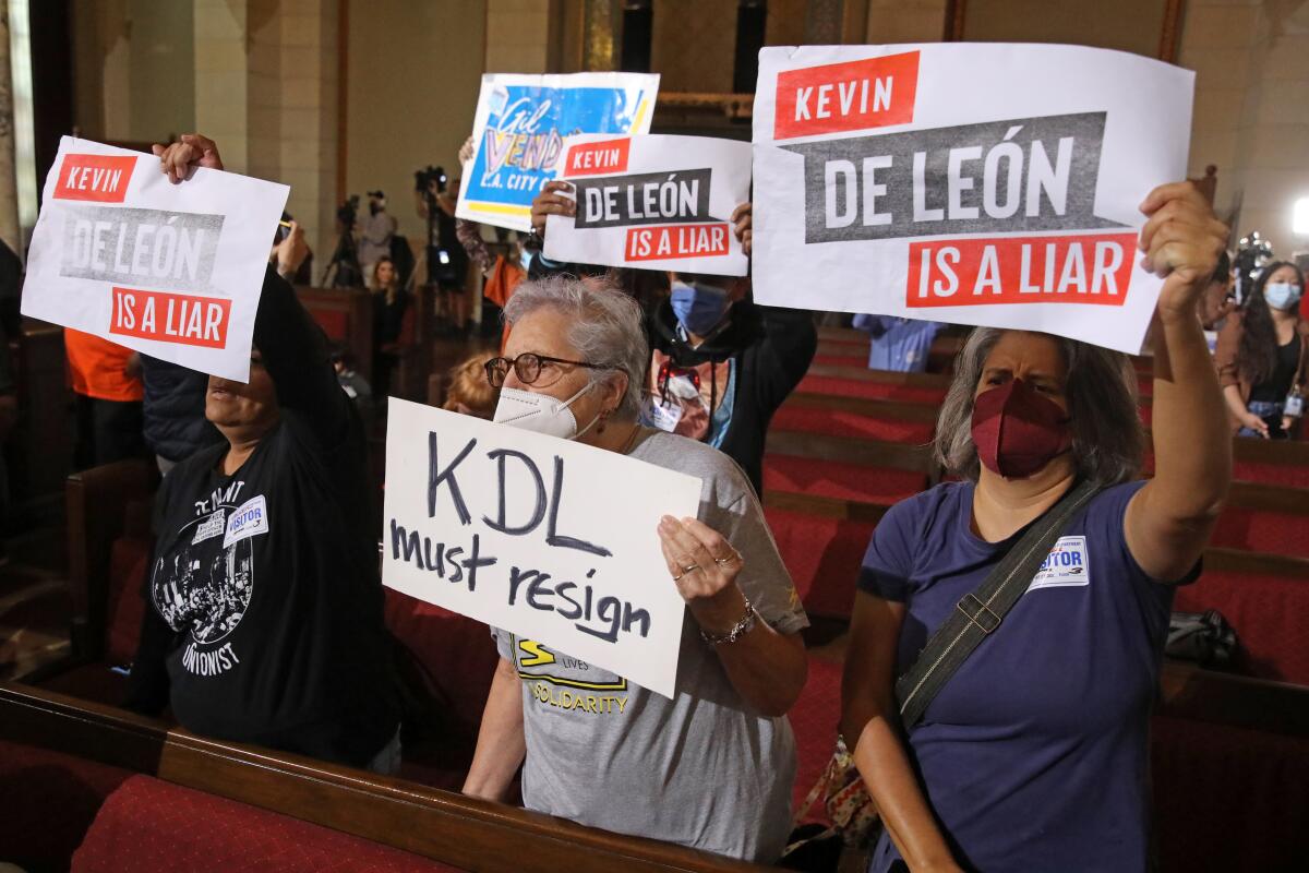 Protesters inside council chambers hold signs that say "KDL must resign" and "De Leon is a liar."