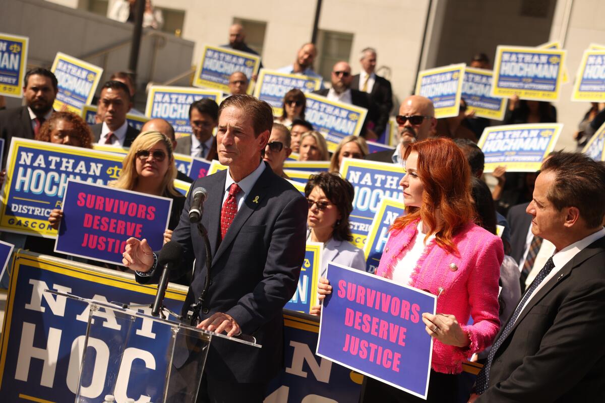 Nathan Hochman speaks into a mic as supporters behind him hold signs with his name or the words "Survivors deserve justice."