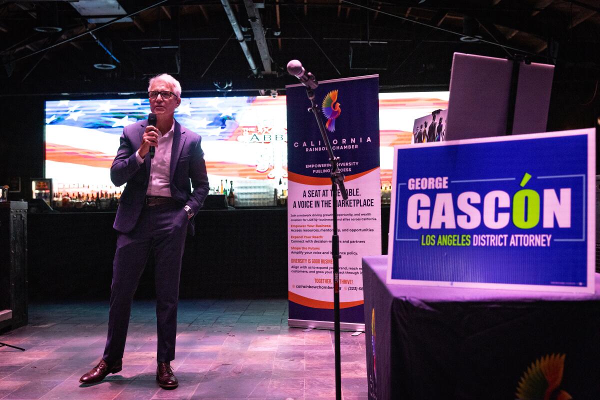 George Gascón stands in front of a large screen bearing a stylized American flag, speaking into a mic beside campaign signs