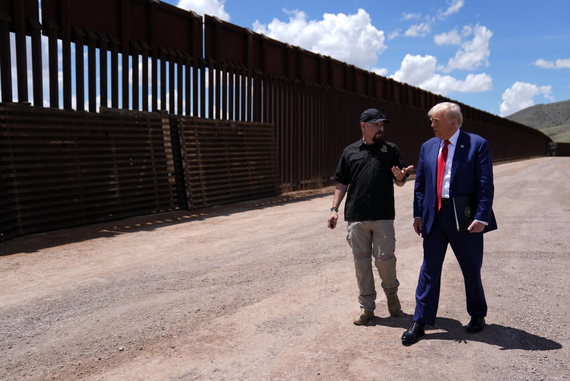 Two men stand near a border wall. 