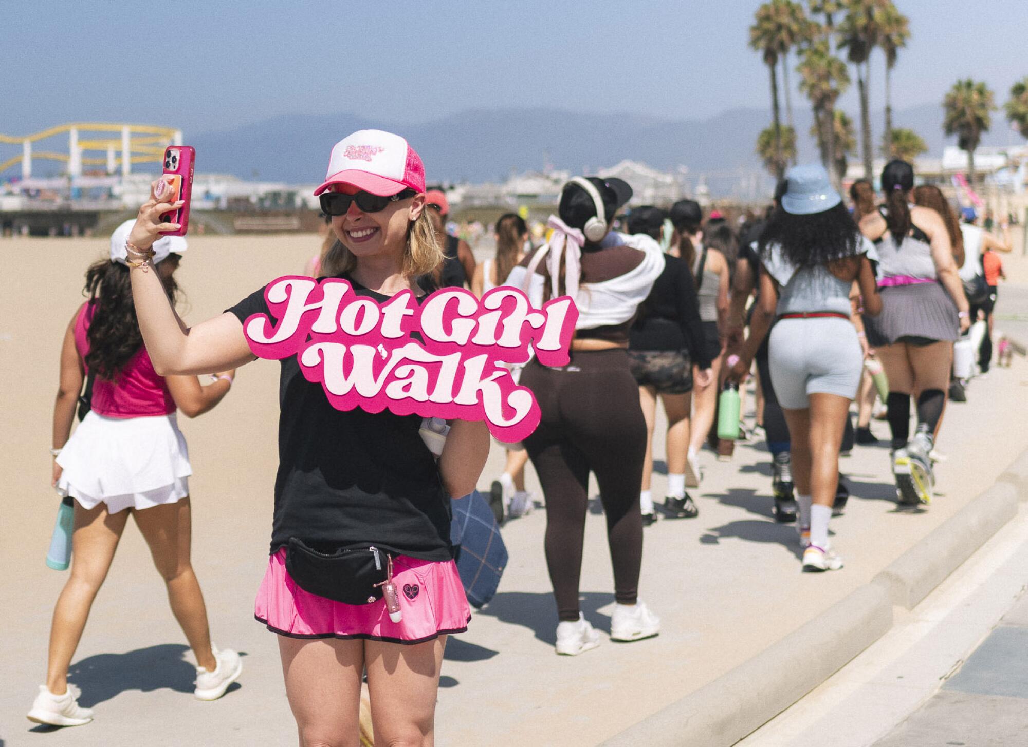 A woman holding a Hot Girl Walk sign directs people along a beachfront path