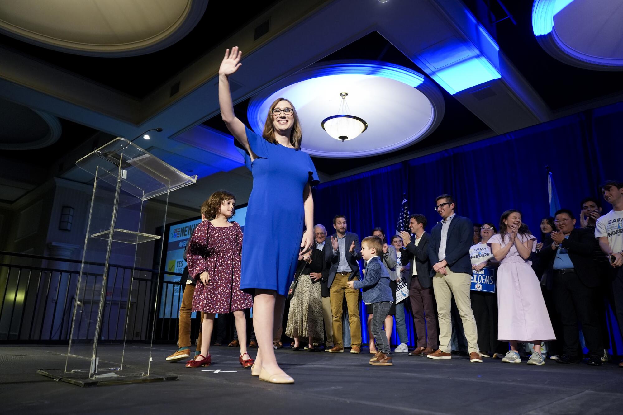 Sarah McBride stands and waves to off-camera supporters; behind her, children dance and more supporters applaud