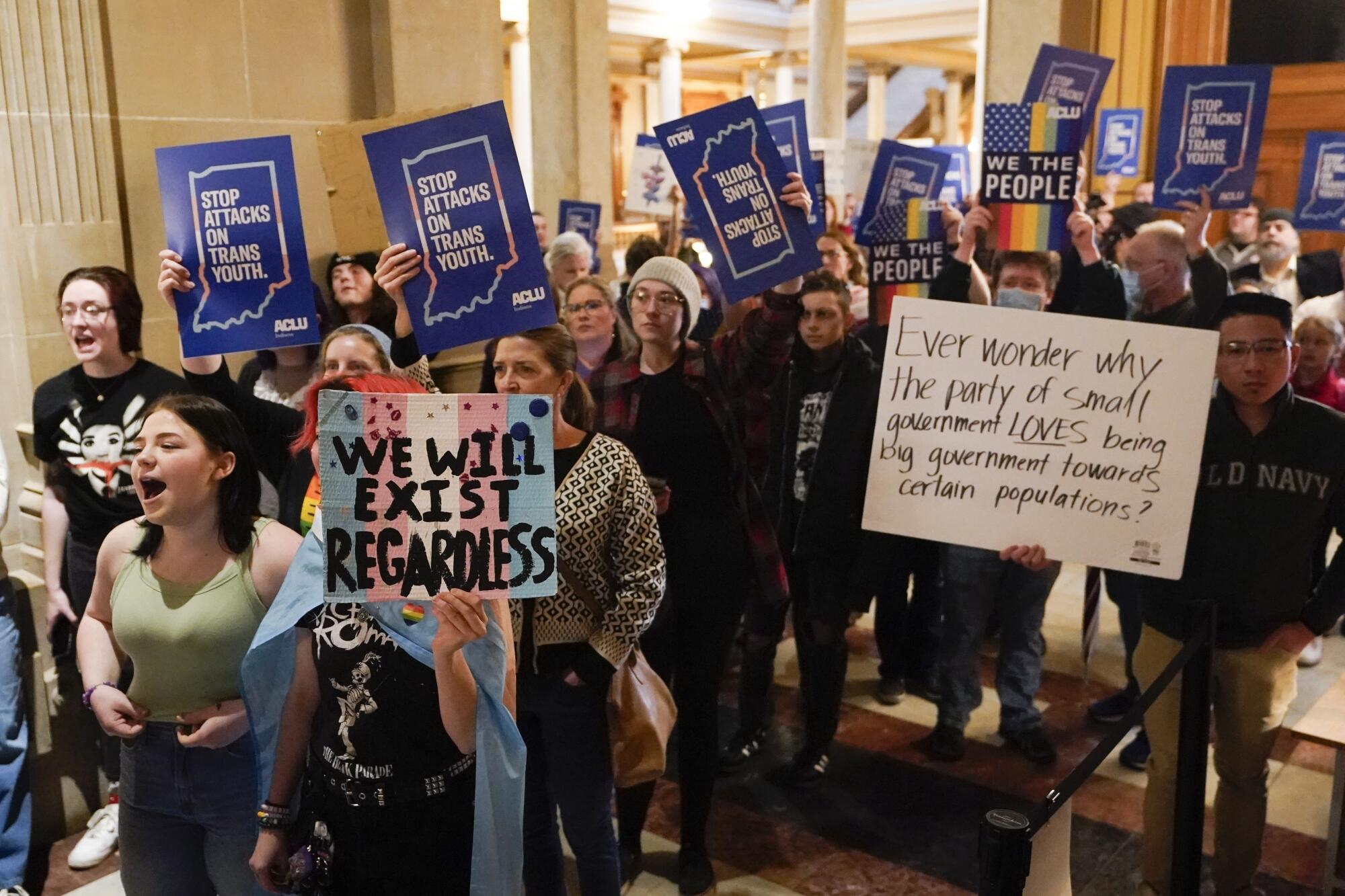 A crowd inside a building chants, holding signs with messages including "Stop attacks on trans youth" and "We the people"