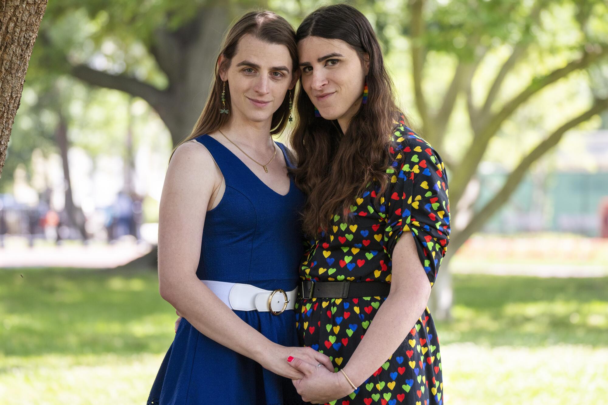 Zooey Zephyr and Erin Reed lean into each other and hold hands for a photo in a parklike setting with large trees 