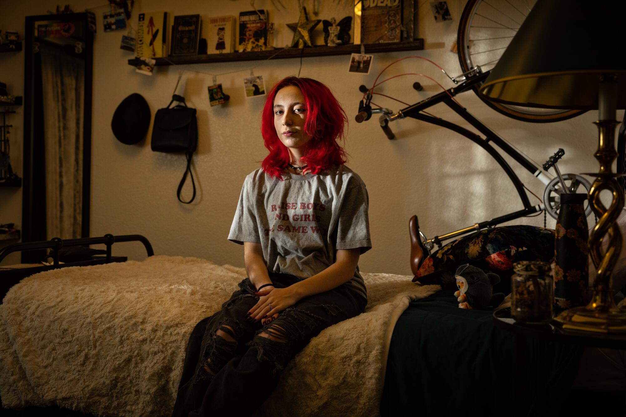 Milo Easley sits on a bed in a dim room, wearing a T-shirt that reads "Raise boys and girls the same way"