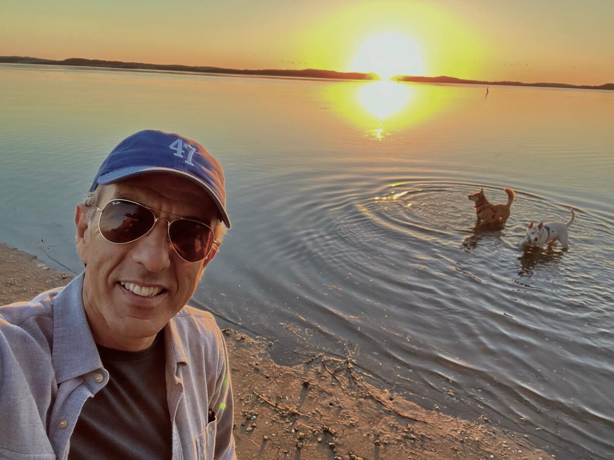 A man with sunglasses and a cap with the number 47 stands on a beach with a dog in the water during sunset