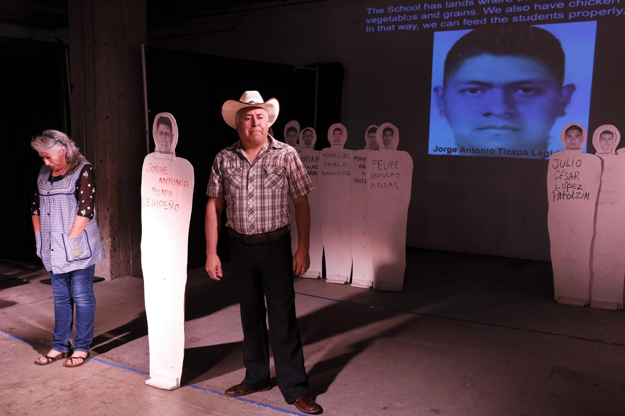 A woman and a man onstage among cardboard cutouts representing missing students. A man's face is projected on the wall.