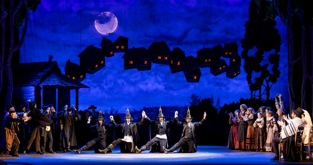 Four men dance in traditional Russian style as other actors watch in "Fiddler on the Roof"