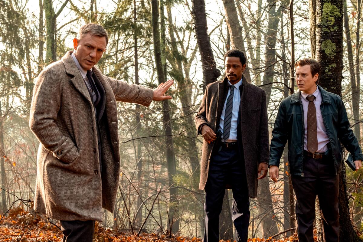 Three men stand in the woods, one holding out his hand to keep the other two back.