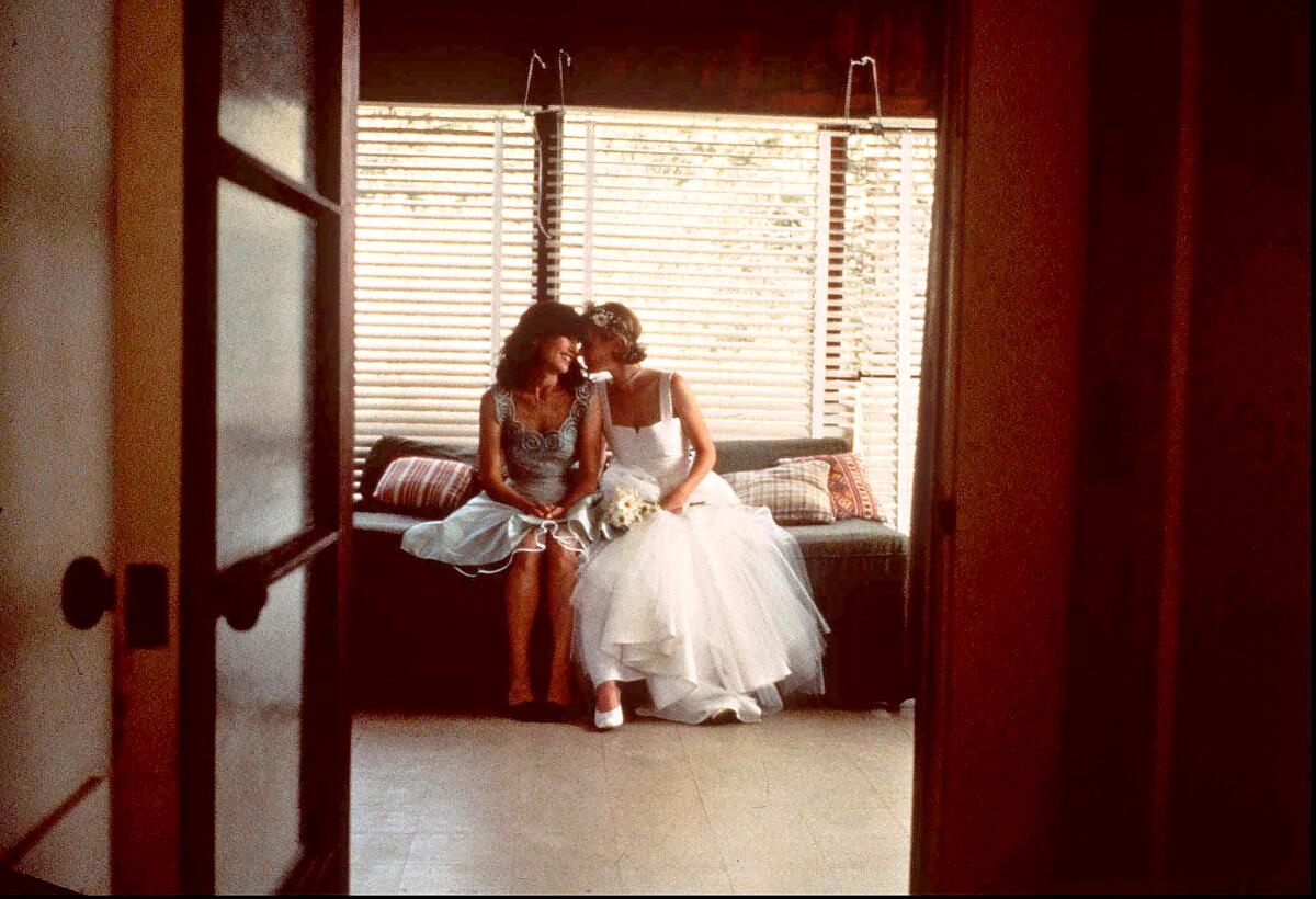 Two women, one in a bridal gown, sit on a couch and speak close together.