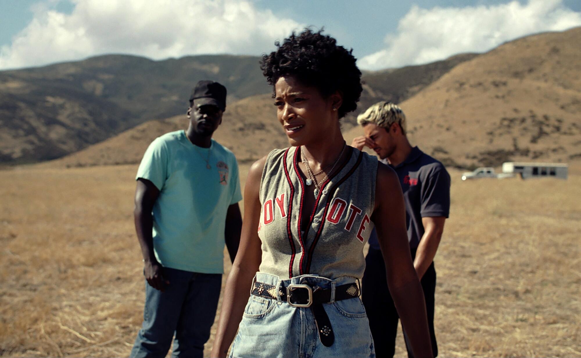 In a huge field, a woman stands, looking a little frightened, with two men standing behind her.