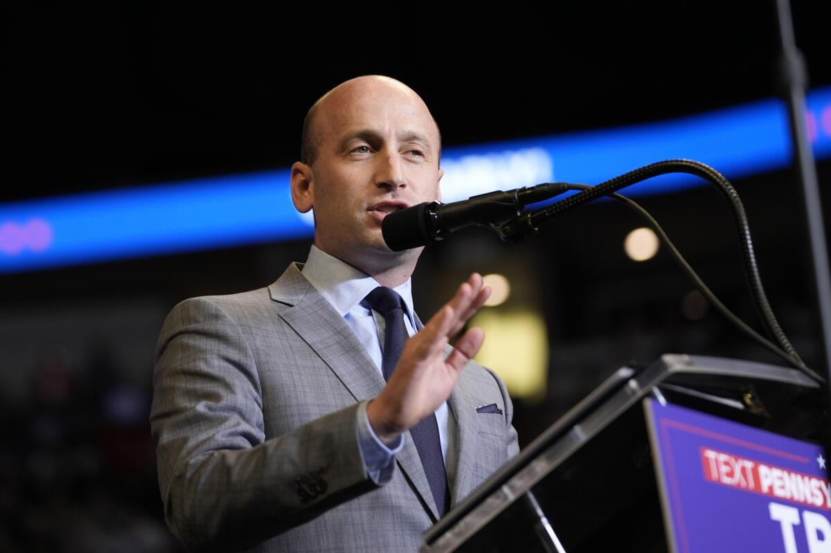 A bald man, in gray suit and dark tie, gestures with his hand while speaking at a microphone