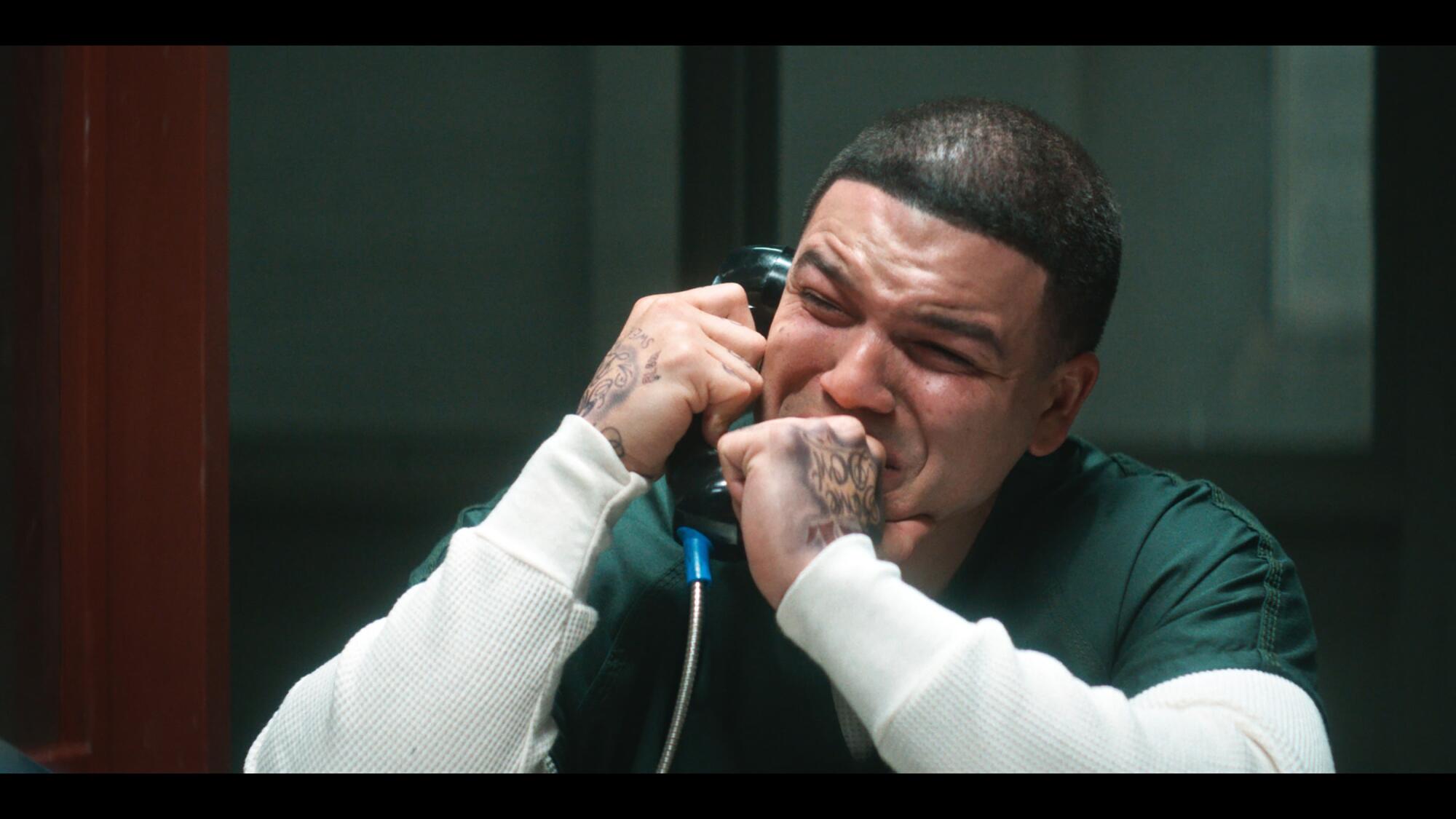 A crying man in a green prison jumpsuit and white shirt holding a telephone receiver to his ear.