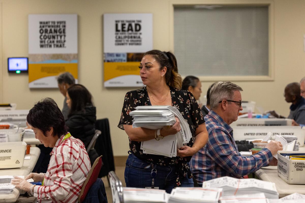 A woman stands holding a stack of papers as people seated on either side of her work at tables  