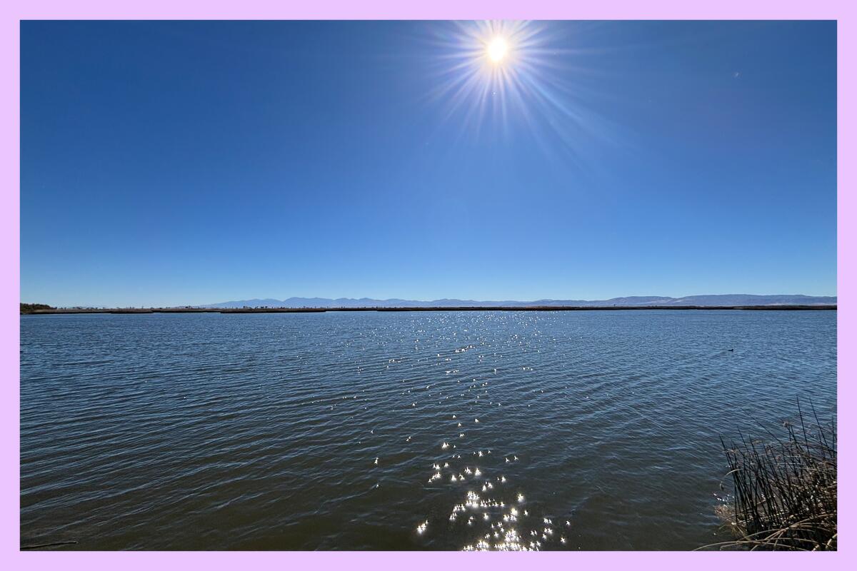 A large body of blue water with a shining sun and a mountain range in the distance