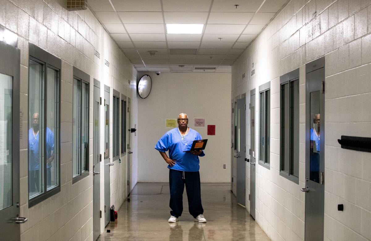 A man in blue prison garb standing near the end of a concrete block hallway, holding a laptop