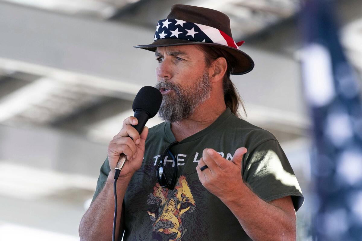 A bearded man in a hat with a flag bandanna around it speaks while holding a microphone