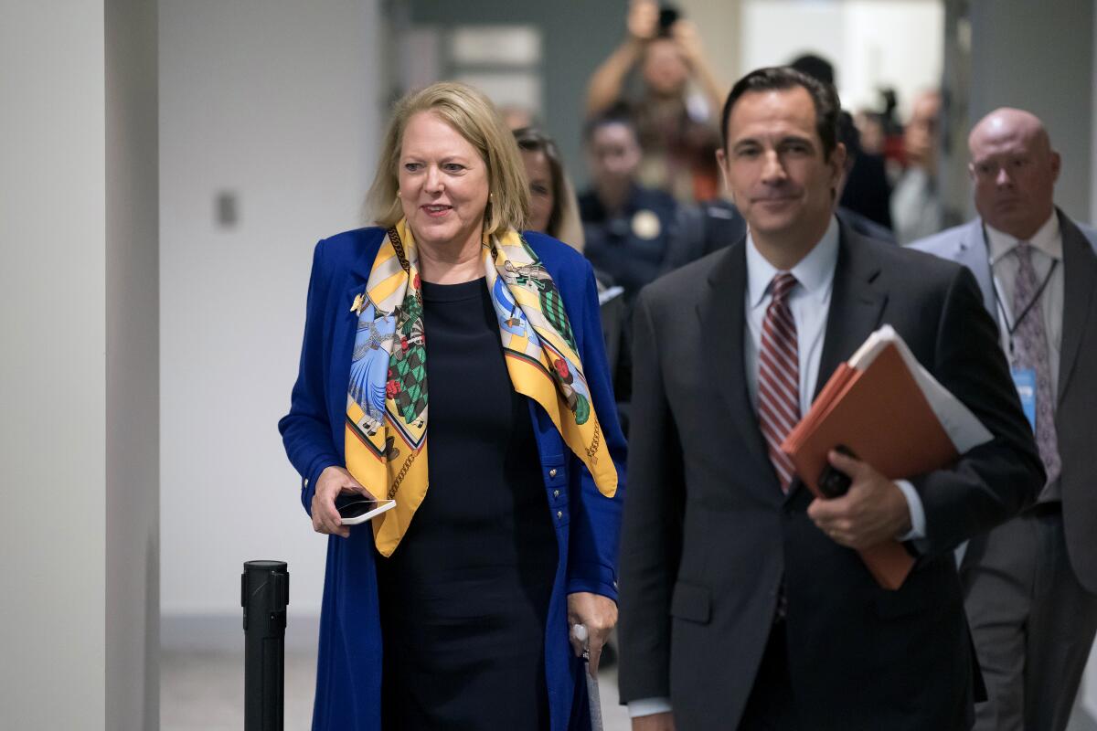 A woman with a blond bob haircut, in a blue coat and colorful yellow scarf, walks behind a man in a suit and striped tie 