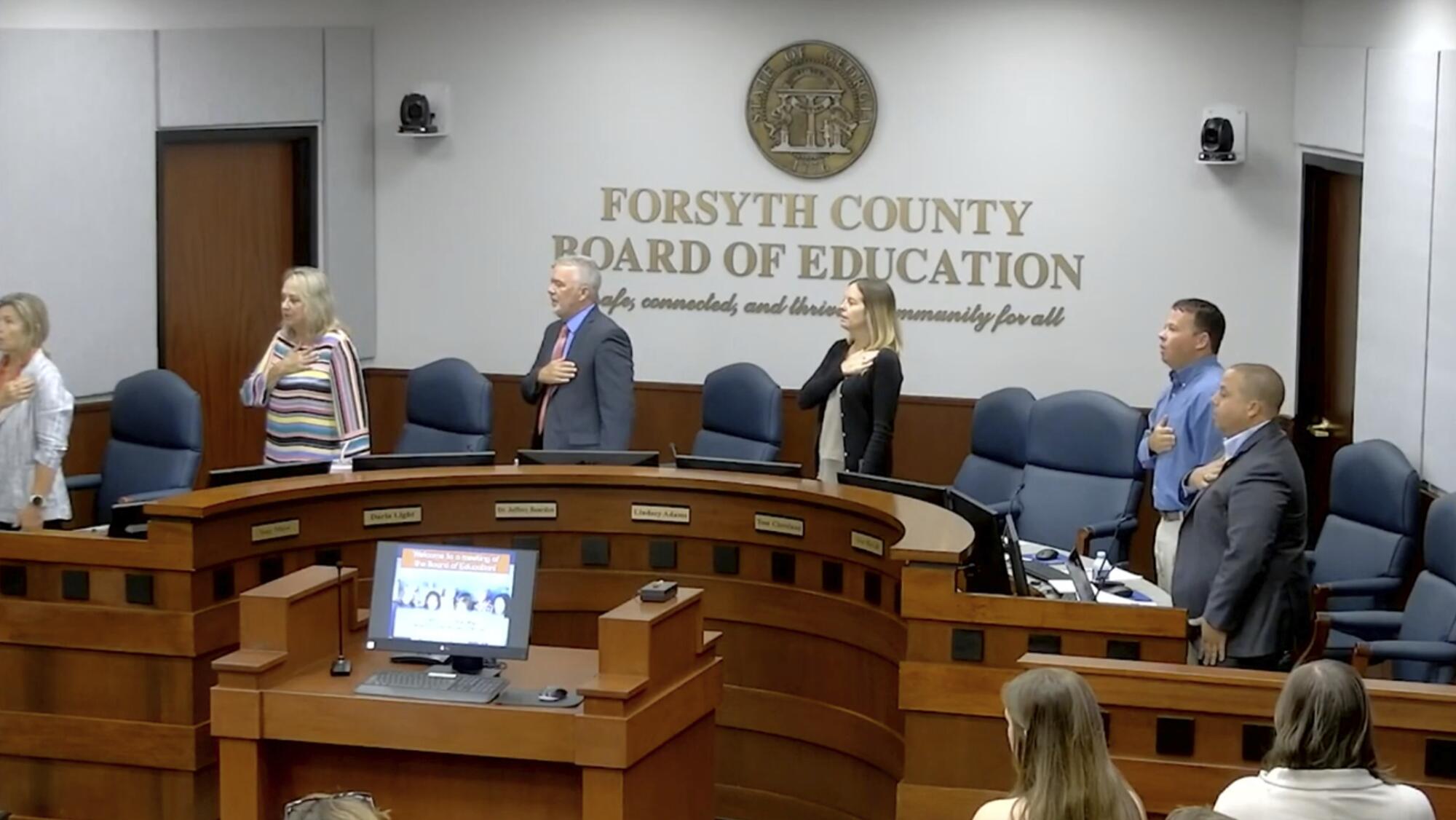 Board members stand at the dais for the Pledge of Allegiance