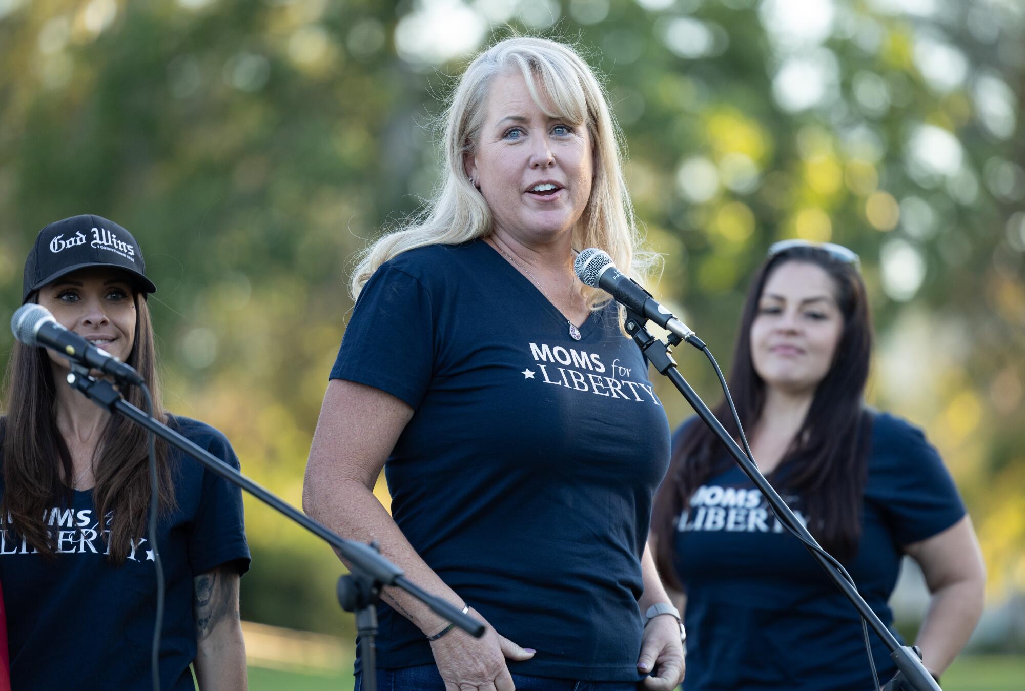 A woman with a "Moms for Liberty" shirt speaks into a microphone as others watch 