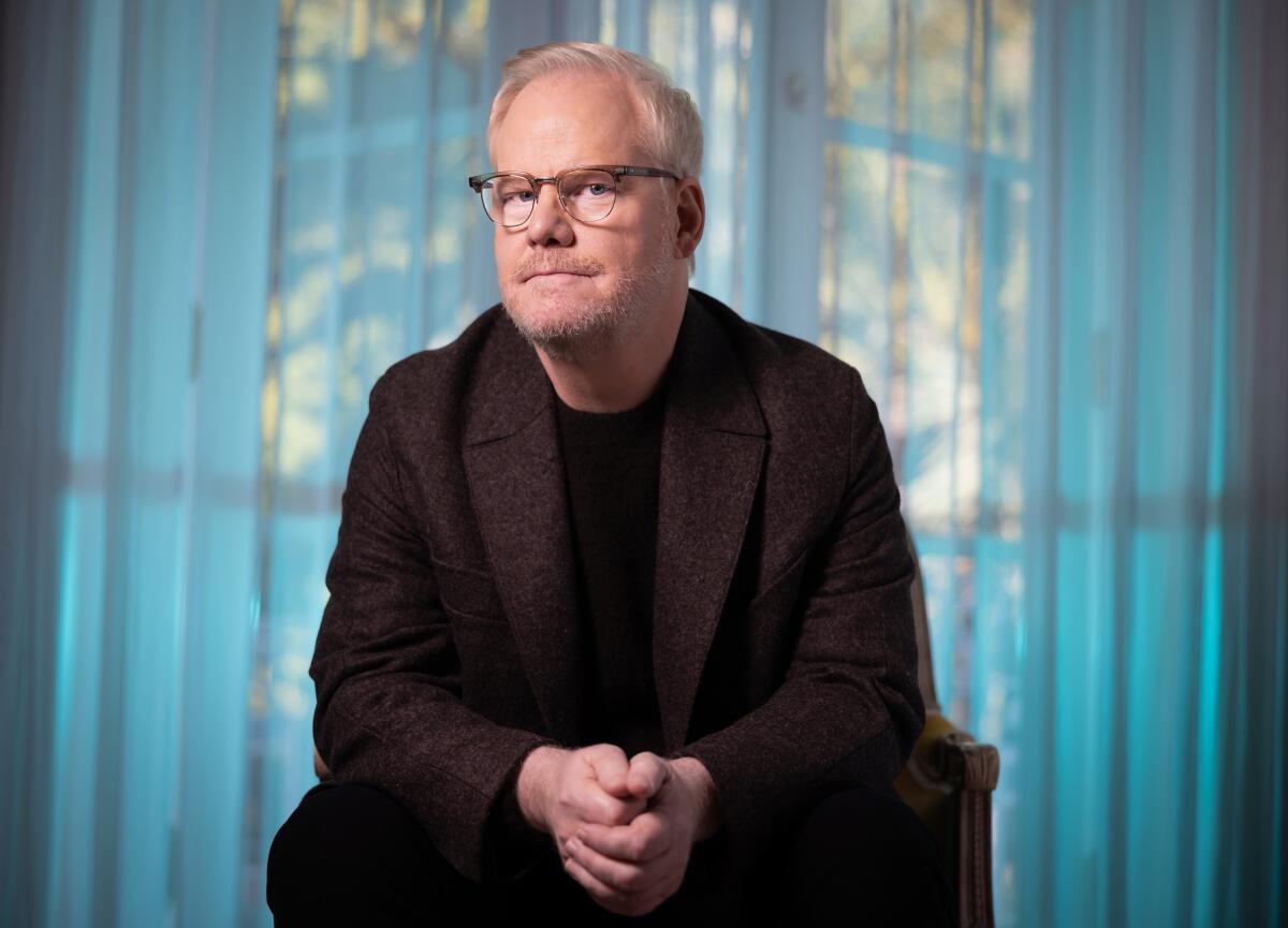 Man sitting in a chair in glasses and a suit in front of a sheer-curtained window