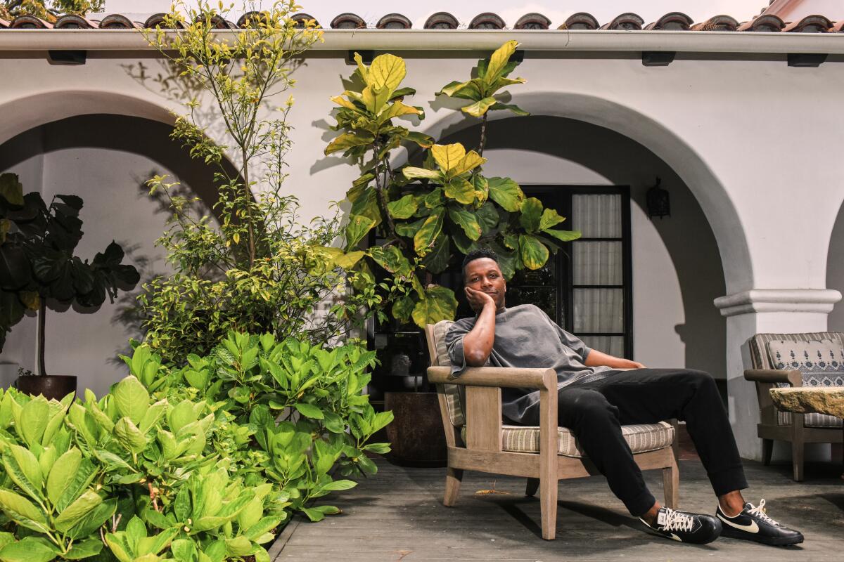  Leslie Odom Jr. sits outside a building's arched windows with shrubs behind him