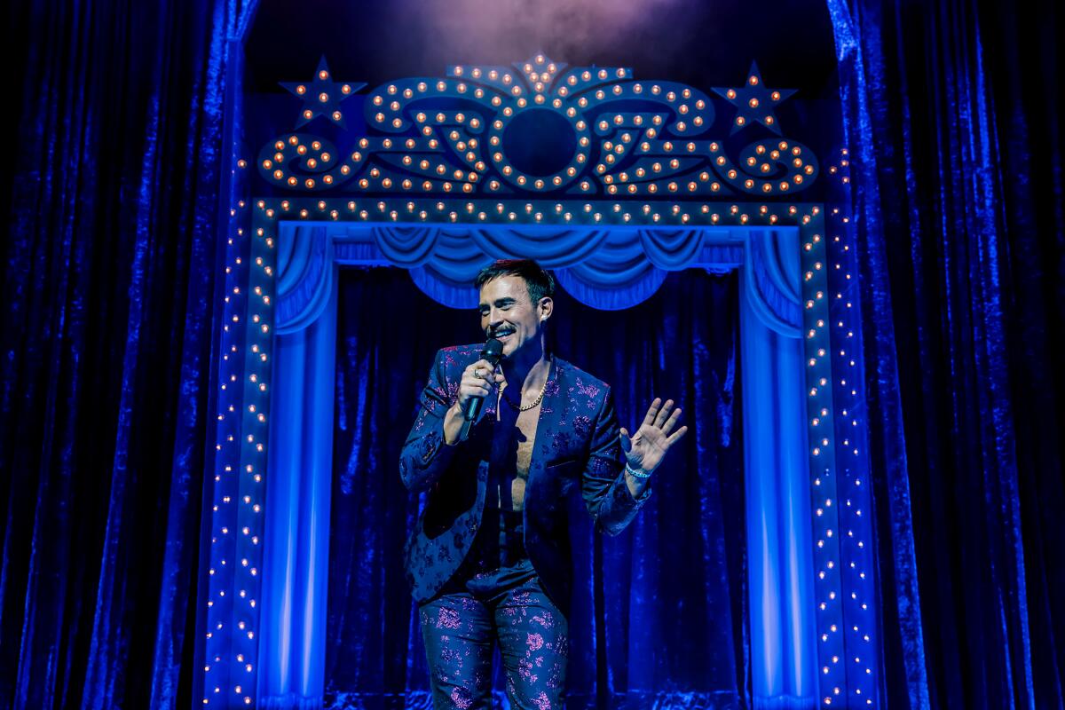 A man in a blue suit gestures and sings into a microphone, in front of an elaborate proscenium arch