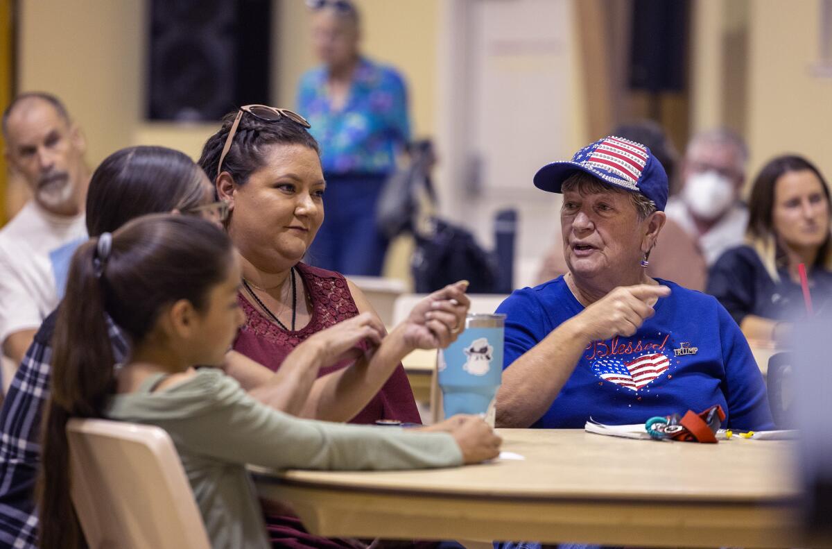 A woman in a blue cap and shirt with American flags points as she talks to her neighbors at one of several tables of people