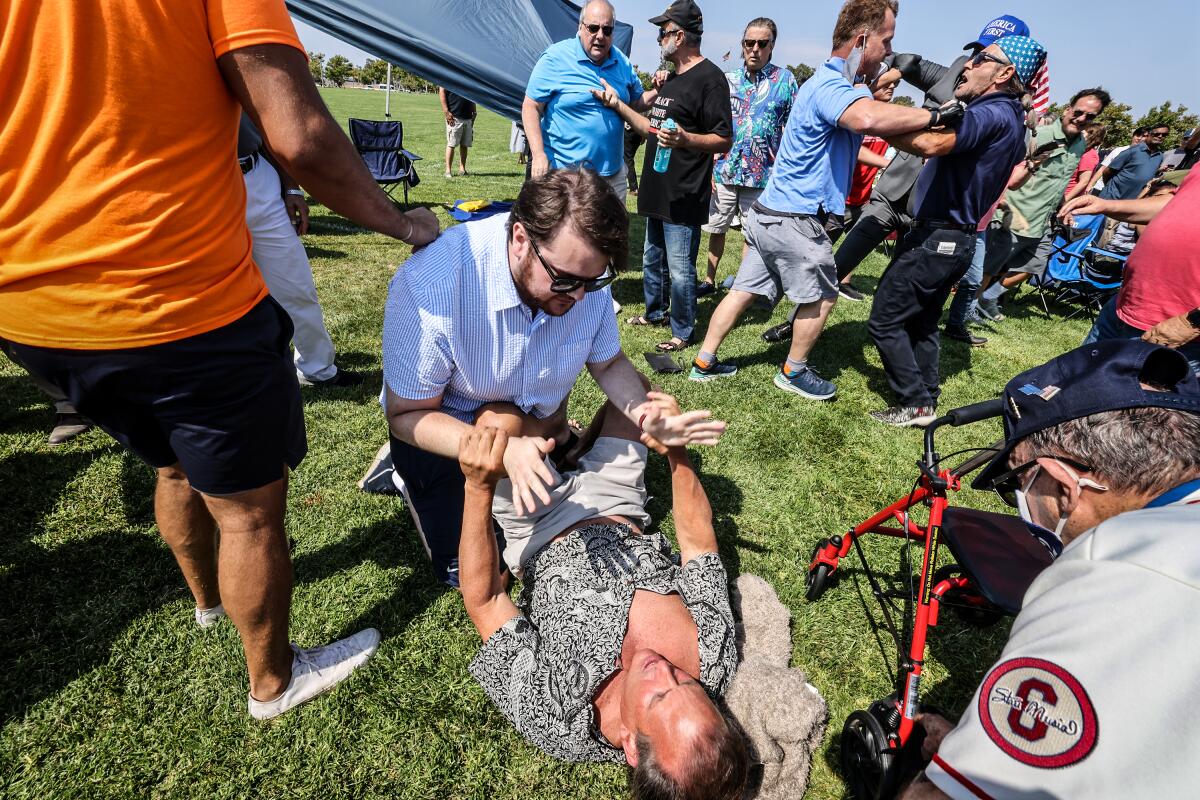 In a crowd, a man lying on the ground grips the wrists of another man. Two men shove each other in the background. 