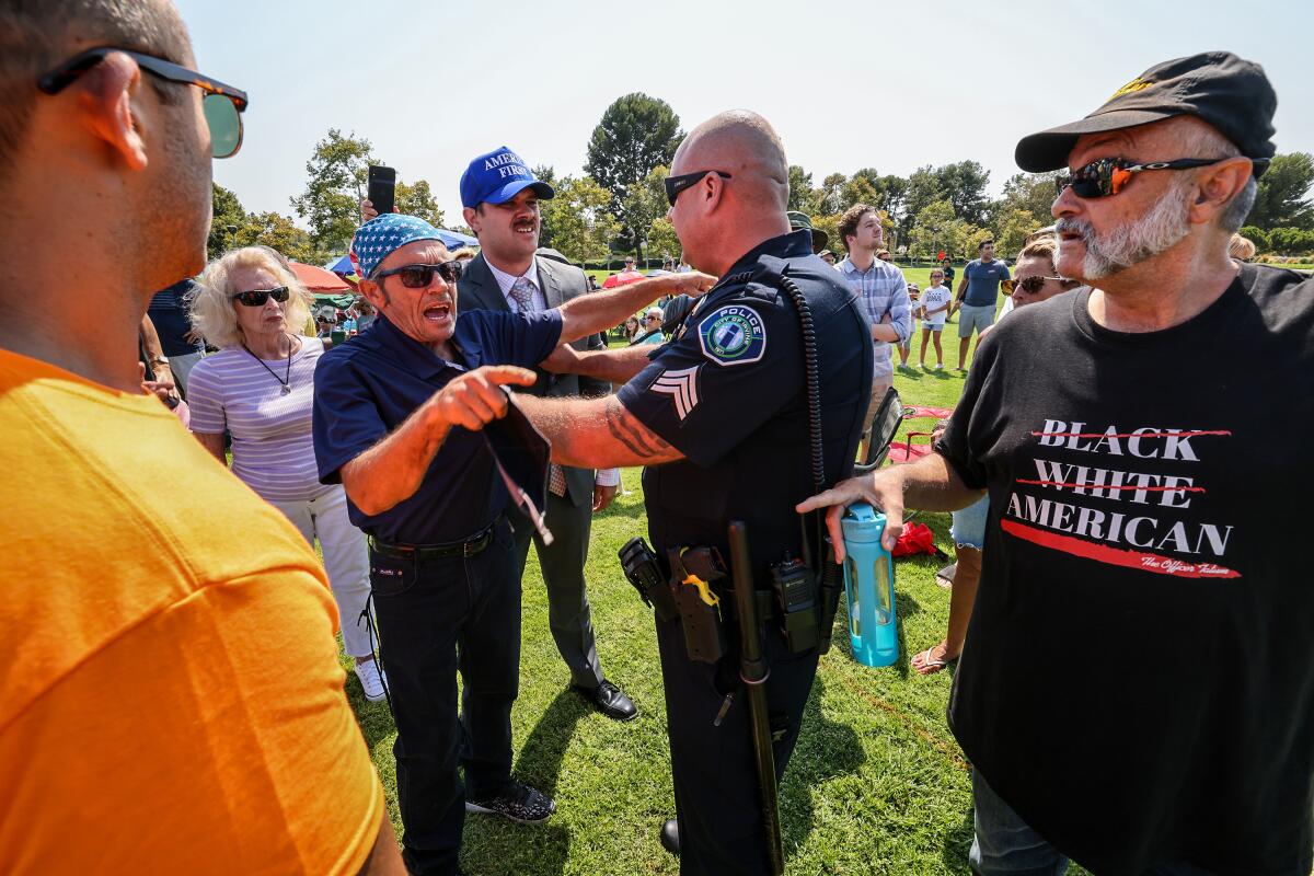 A man in a flag head covering is held back by a police officer as he yells and points at another man.