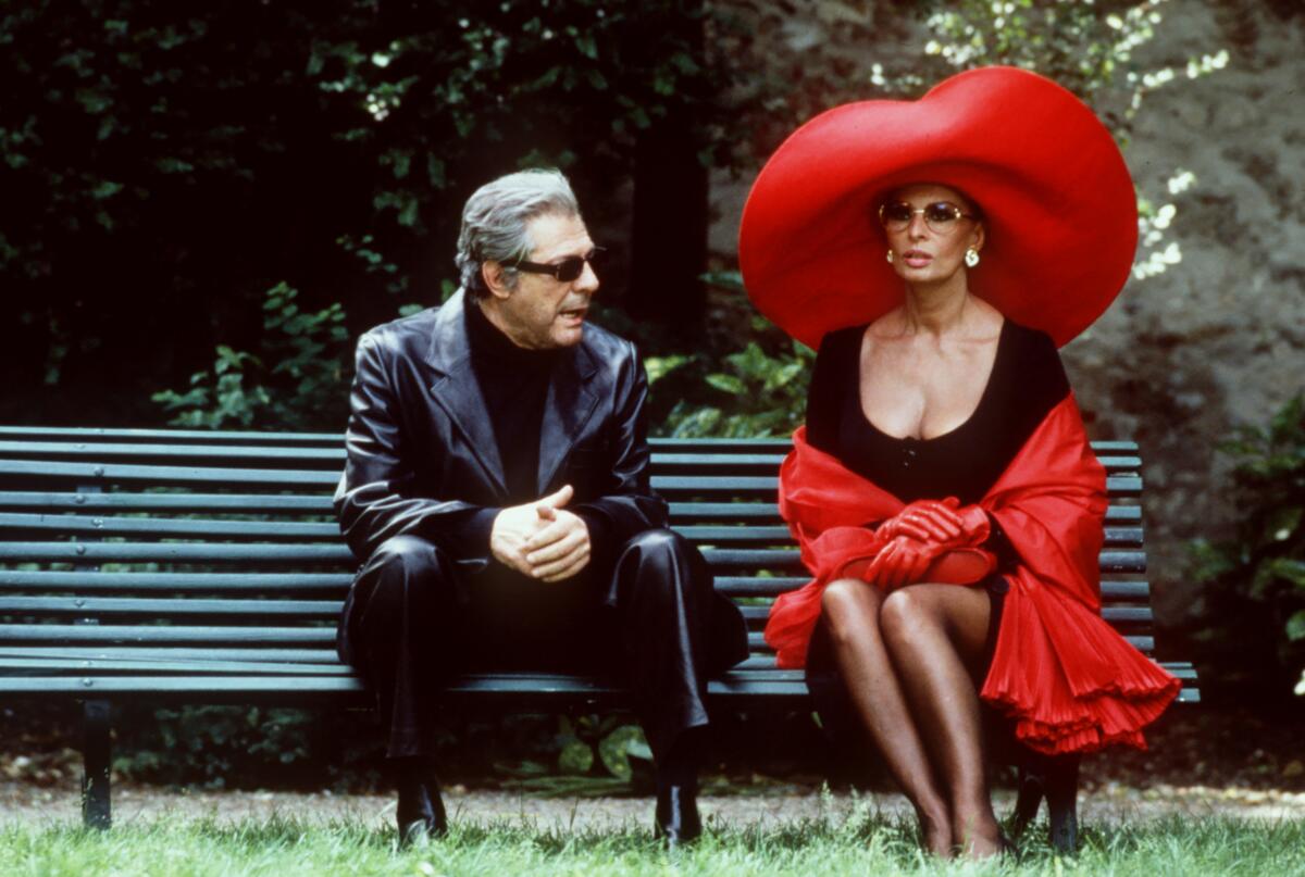 A man in shades sits on a park bench with a glamorous woman.