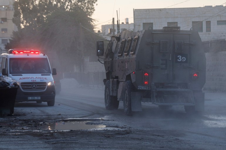 a large military vehicle in front of an ambulance on a dusty street