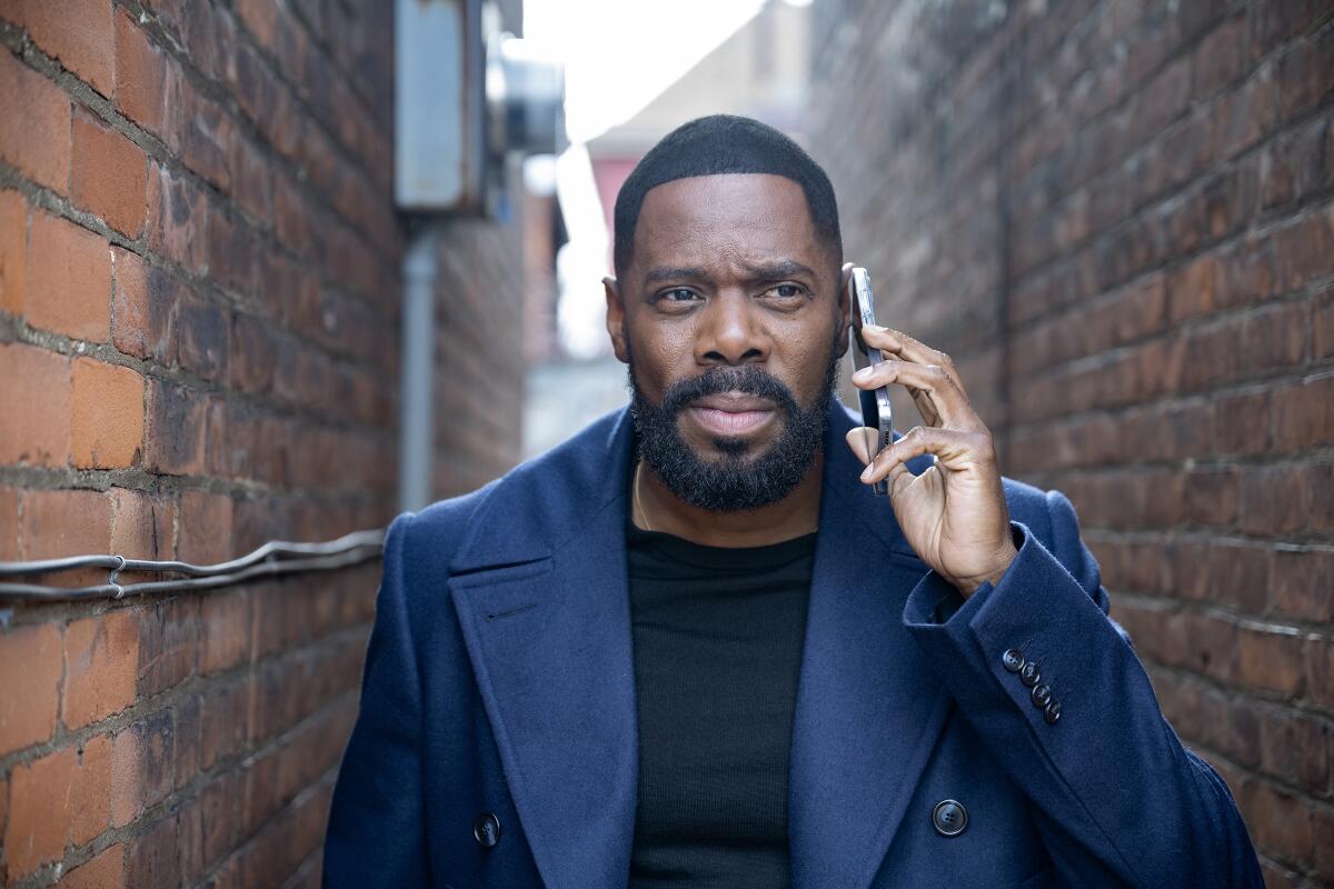 A man in a blue peacoat and black shirt stands in an alleyway with a phone held to his ear.