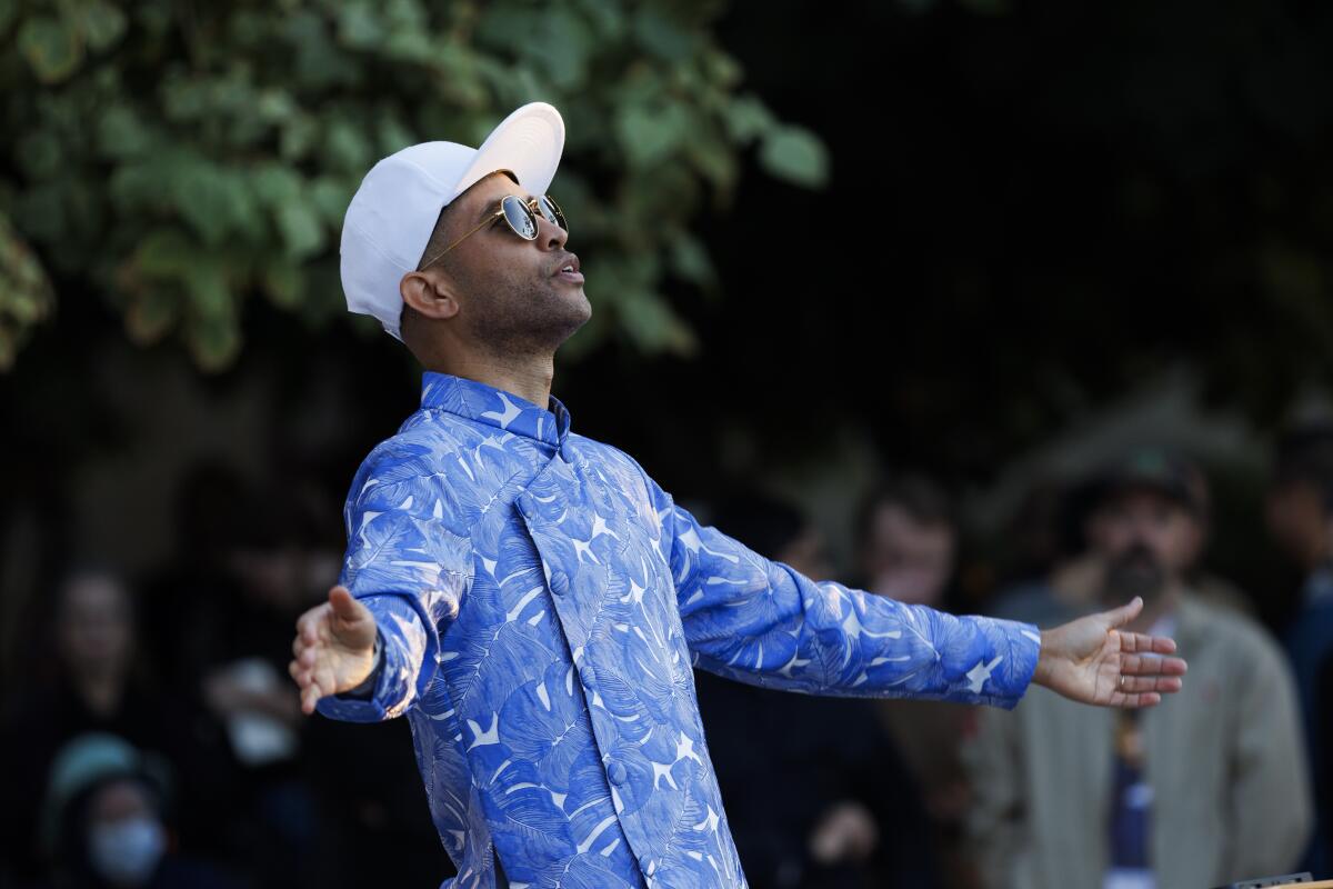 A man in blue shirt, white cap and sunglasses conducts an ensemble in the garden behind Disney Hall.