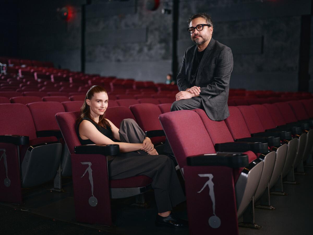 A woman in a black tank top and a man in eyeglasses sit in an empty theater.