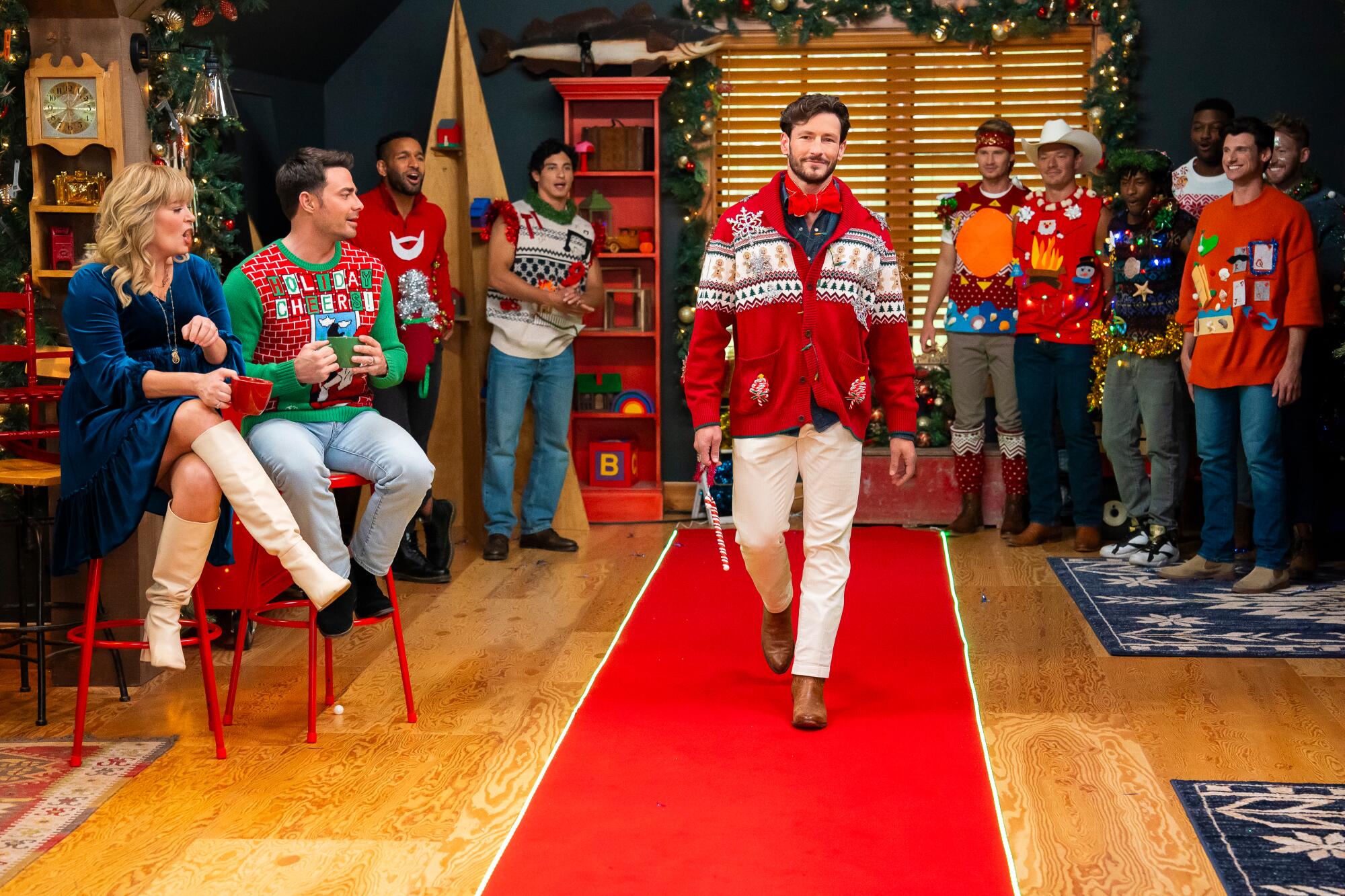 A man in a Christmas sweater walks down a red carpet as people watch.