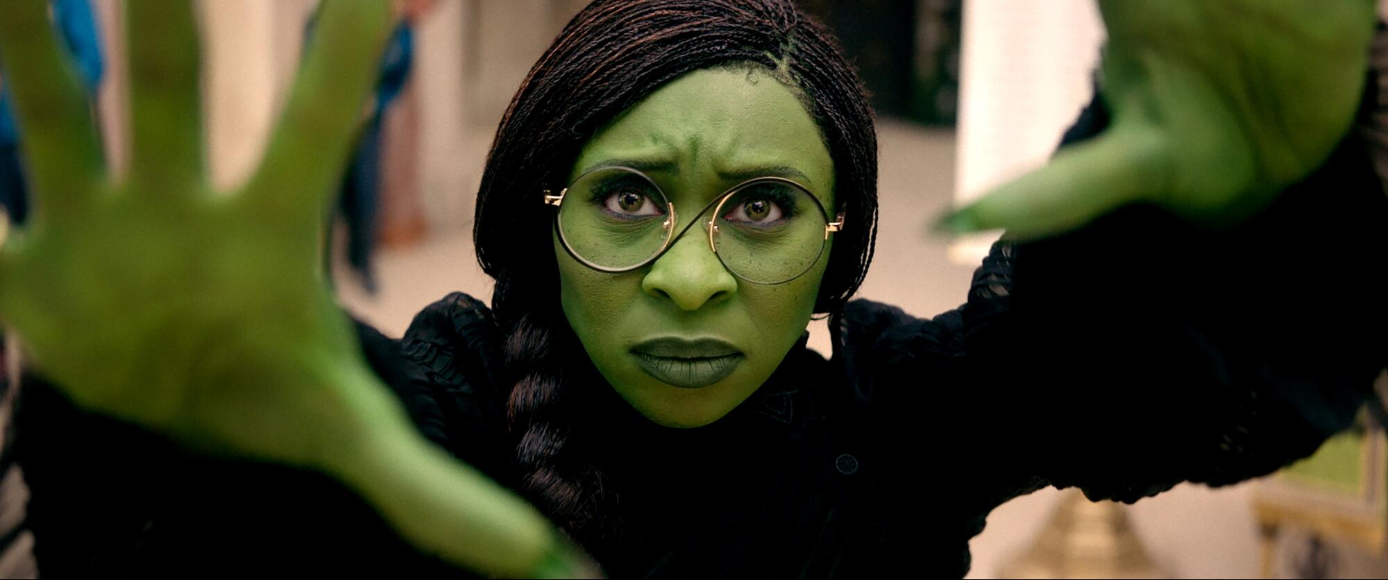 A woman with green hands and face stares ahead in a scene from "Wicked."