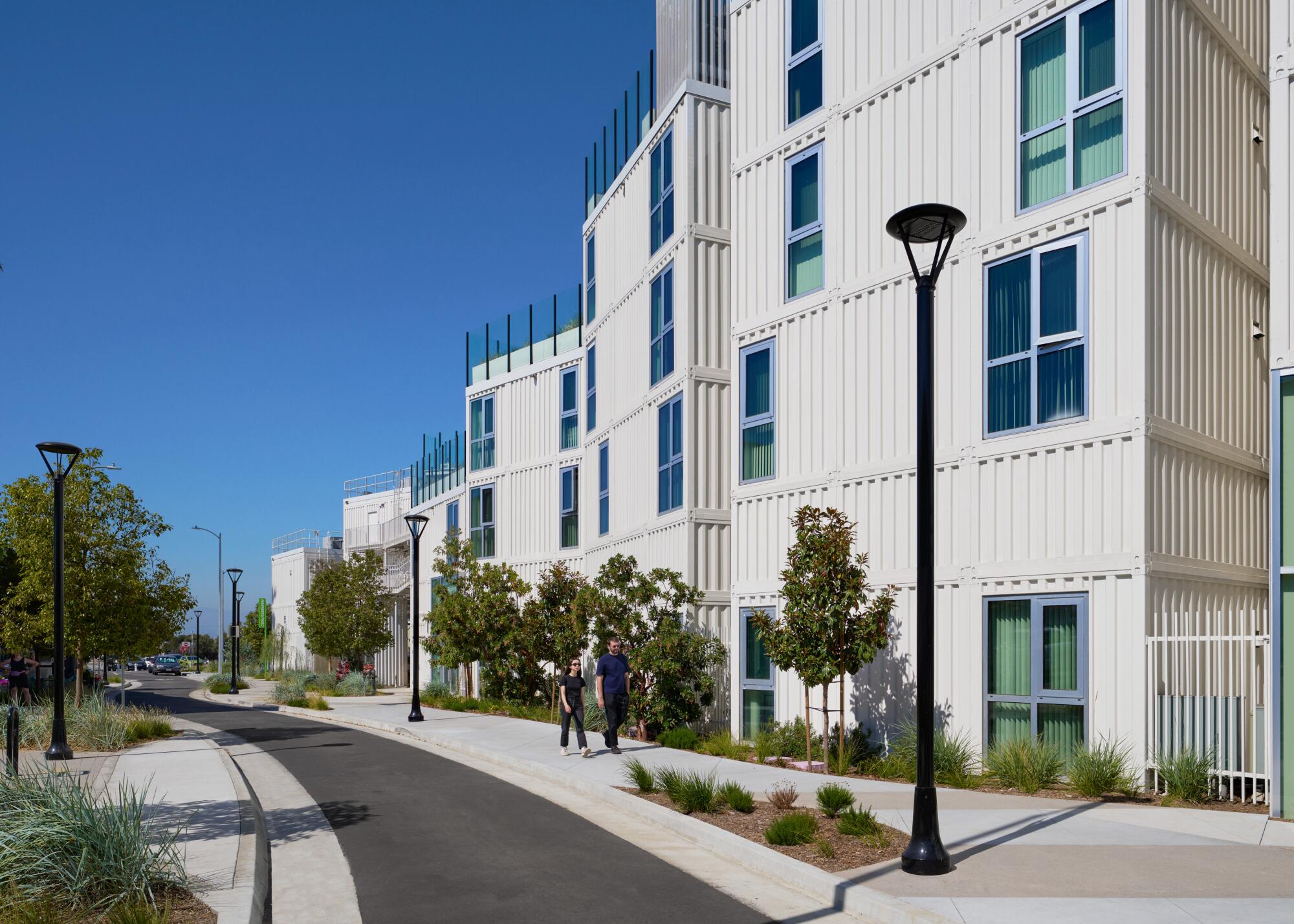 Two people walk on a sidewalk next to three- and four-story apartments made from shipping containers.