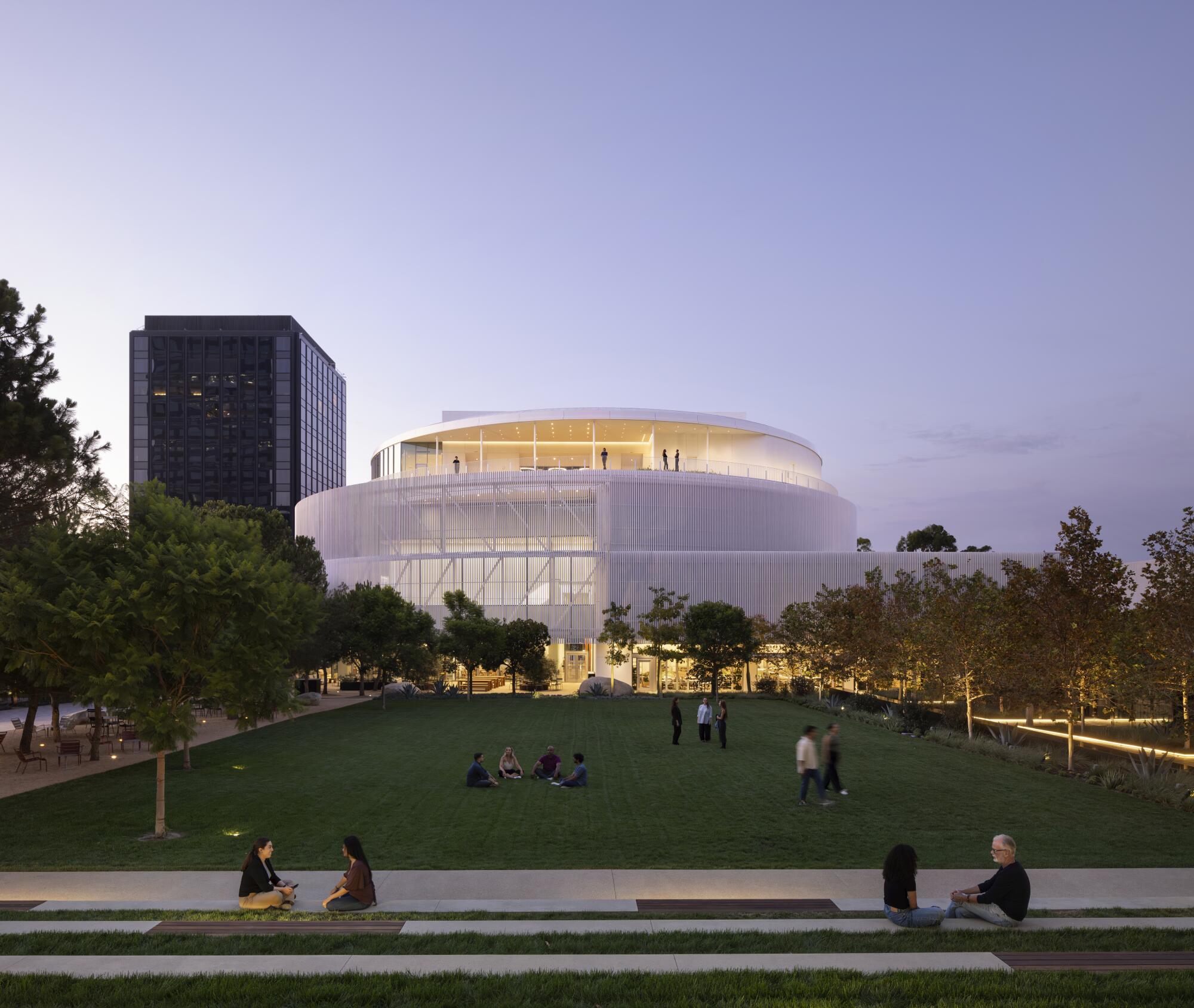 A circular white building rises above a tree-lined grassy field. 