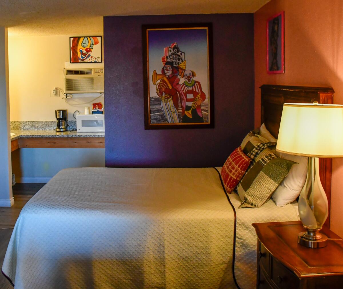 Artwork portraying clowns is hung on a wall next to a bed in a motel room.