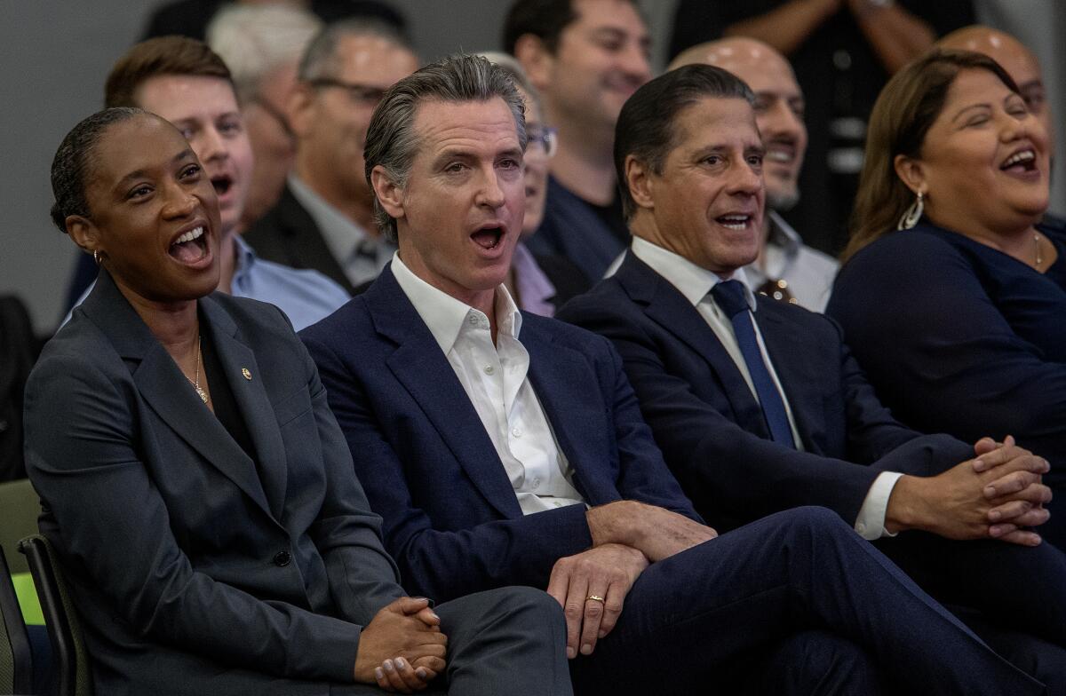 Laphonza Butler and Gavin Newsom sitting in the front row of an audience, chanting along with others