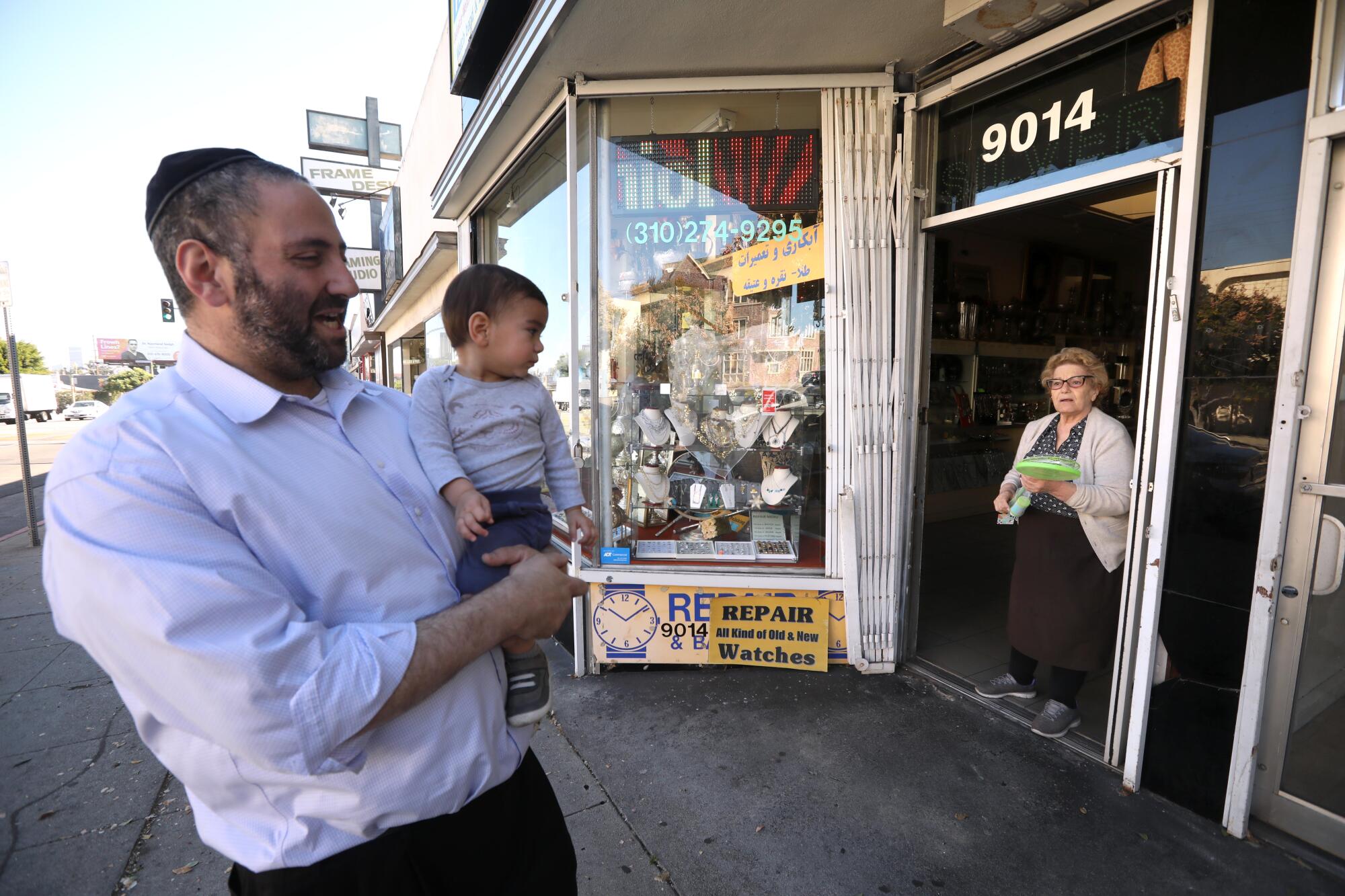 A man holds a boy while both look at a woman in the doorway of a shop.