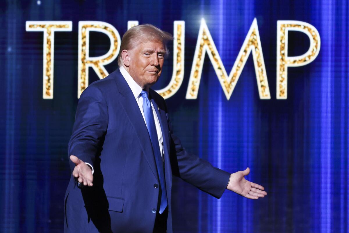 A man in a suit and blue tie holds out his hands in front of a blue background with the word Trump in gold letters