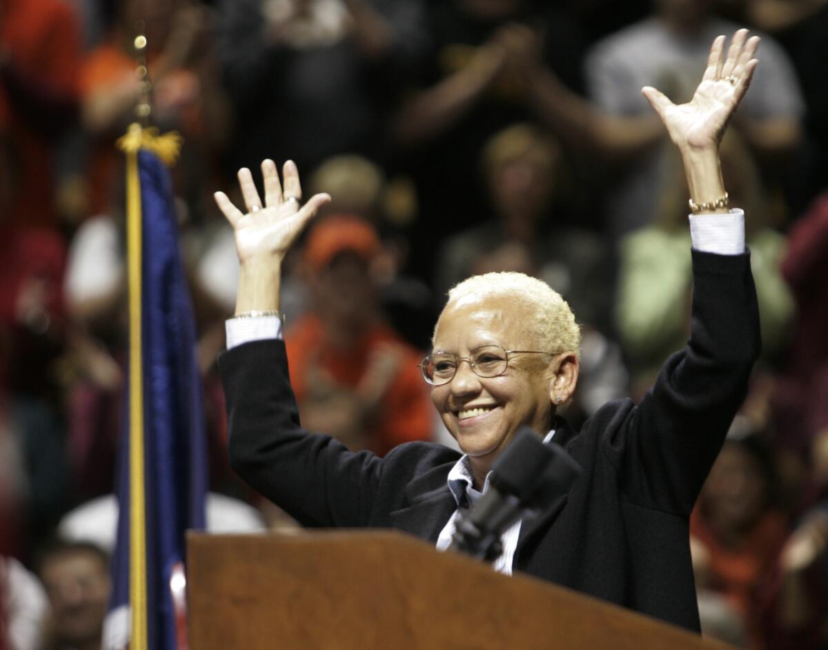 Nikki Giovanni waves both hands in the air while smiling at a lectern in front of a crowd