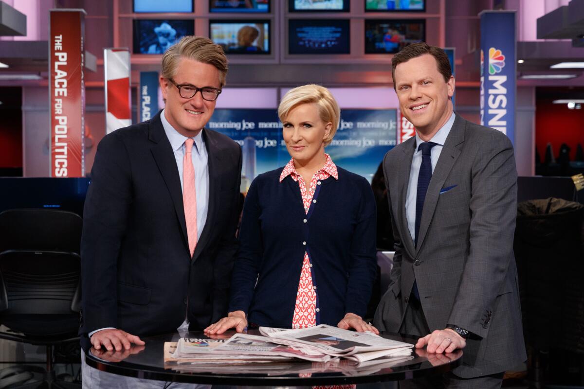 Two men in suits flank a blond woman on a TV show set.