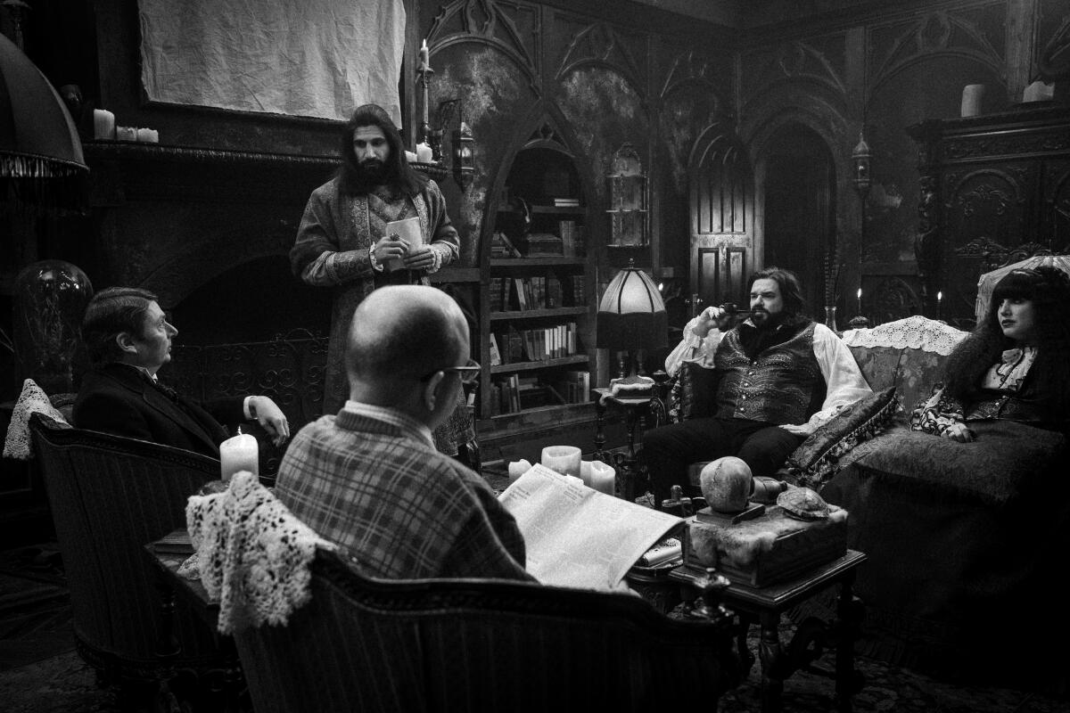 A black and white photo of a man standing at the head of a room as four people sitting in armchairs look up at him.